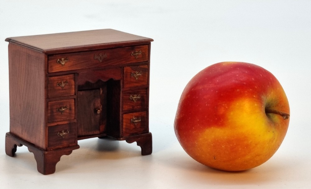 An antique miniature yew wood kneehole desk featuring small drawers, brass handles, and a central cupboard door. View III