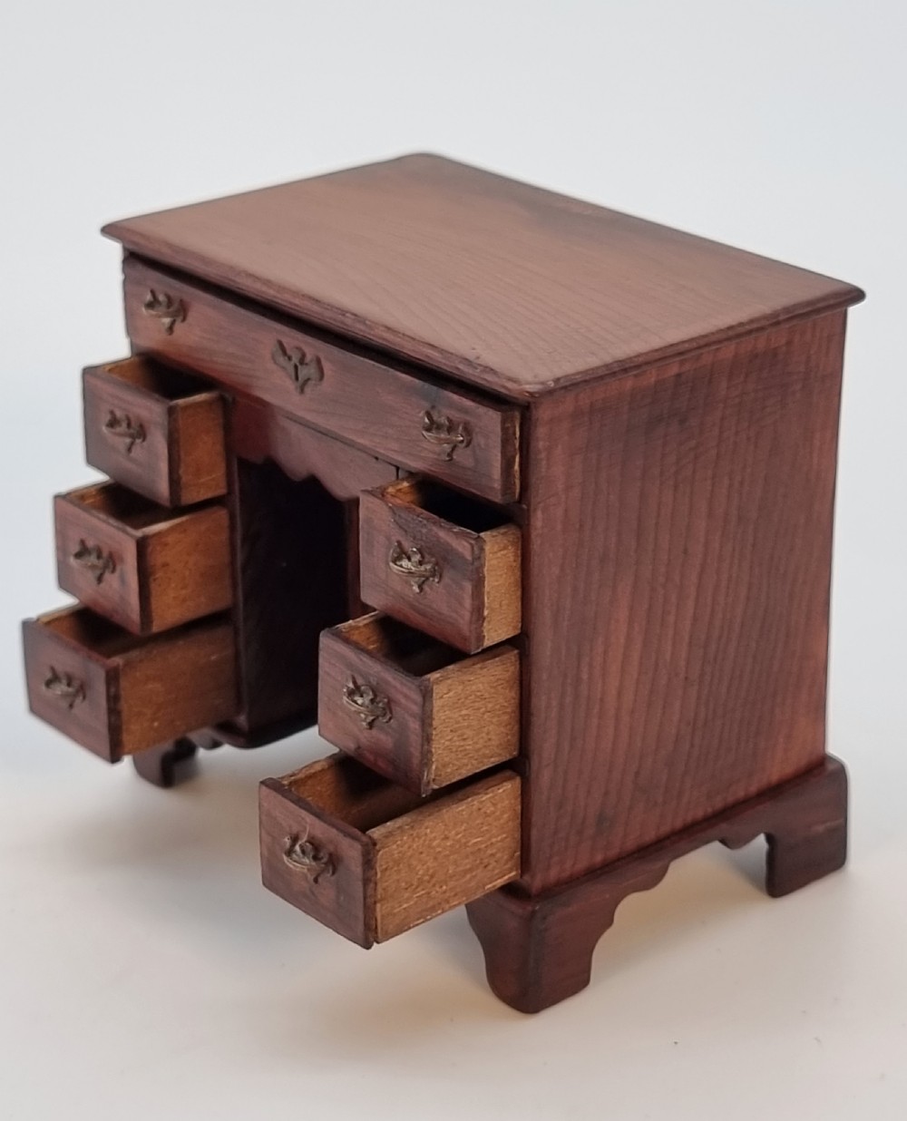 An antique miniature yew wood kneehole desk featuring small drawers, brass handles, and a central cupboard door. View V