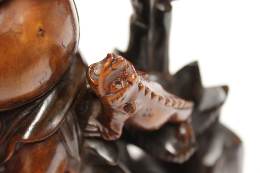 A late nineteenth-century carved brown hardwood figure depicting a seated laughing figure with an open mouth alongside a small mythical creature on a carved rocky base. View XIV