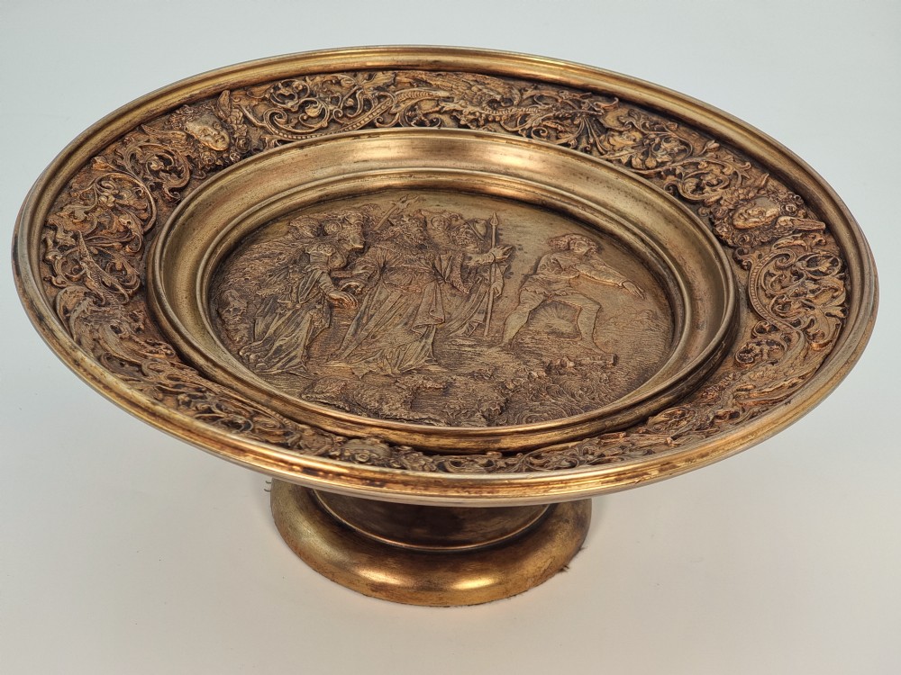 An Aesthetic Movement circular gilt metal tazza on a pedestal foot featuring ornate foliate scrollwork and a central relief medallion.
