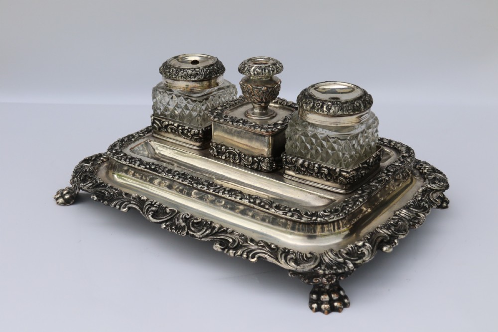 An early nineteenth-century Sheffield plate desk inkstand featuring two cut-glass inkwells and decorative scroll borders raised on paw feet. View V