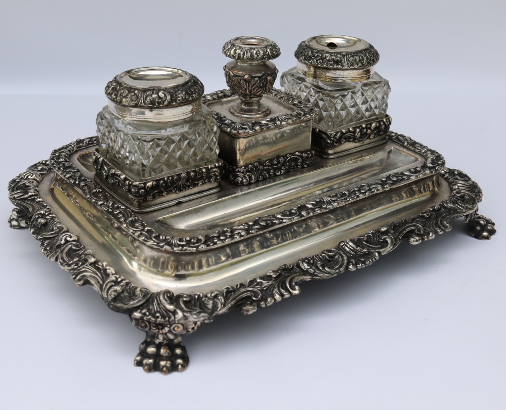 An early nineteenth-century Sheffield plate desk inkstand featuring two cut-glass inkwells and decorative scroll borders raised on paw feet.