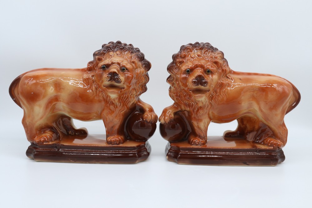 A pair of warm brown glazed earthenware Medici lion figurines standing on integral bases with one front paw resting on a ball.