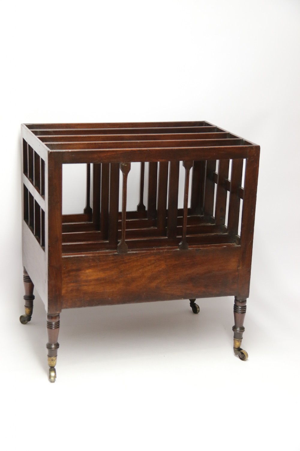 A dark mahogany George III Canterbury featuring slatted vertical partitions, a lower single drawer with brass ring handles, and turned legs resting on brass castors. View III