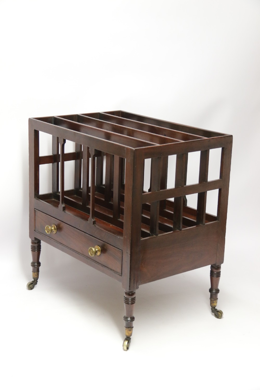 A dark mahogany George III Canterbury featuring slatted vertical partitions, a lower single drawer with brass ring handles, and turned legs resting on brass castors. View V