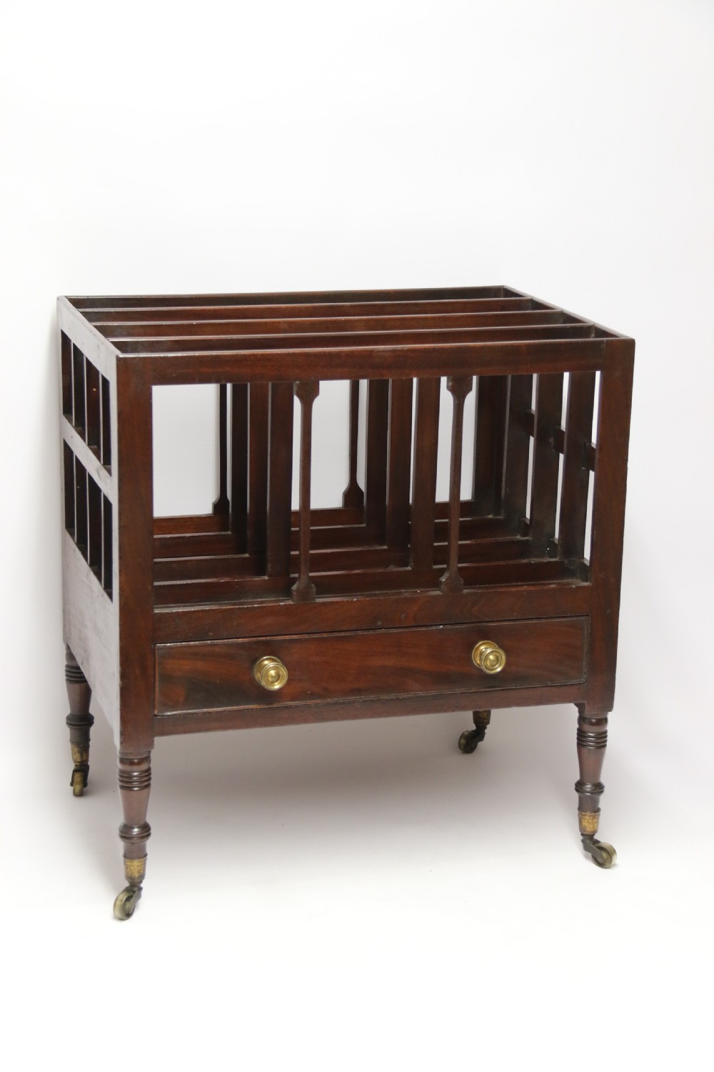 A dark mahogany George III Canterbury featuring slatted vertical partitions, a lower single drawer with brass ring handles, and turned legs resting on brass castors. View VIII