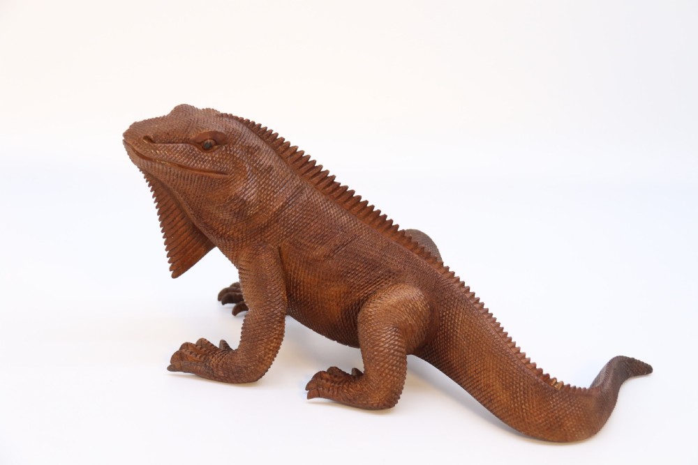 A hand-carved hardwood sculpture of an iguana featuring detailed scale patterns, a serrated dorsal crest, and a prominent dewlap in a warm brown finish. View VII