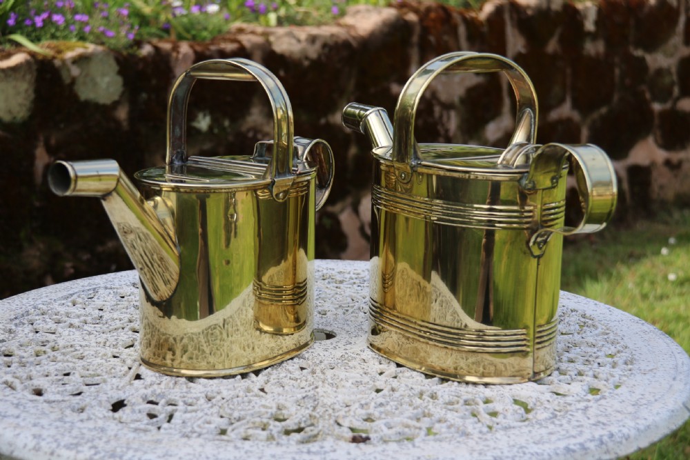A pair of polished 19th-century brass watering cans in differing sizes featuring reeded cylindrical bodies, arched top handles, and angled spouts. View V