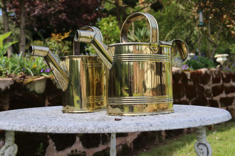 A pair of polished 19th-century brass watering cans in differing sizes featuring reeded cylindrical bodies, arched top handles, and angled spouts. View IX