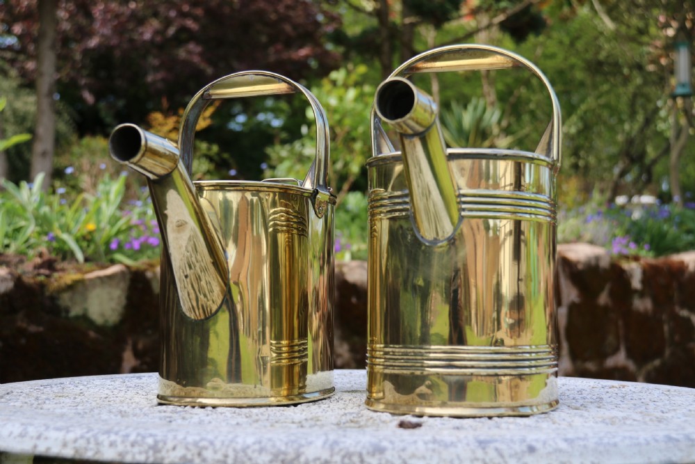 A pair of polished 19th-century brass watering cans in differing sizes featuring reeded cylindrical bodies, arched top handles, and angled spouts. View XIX