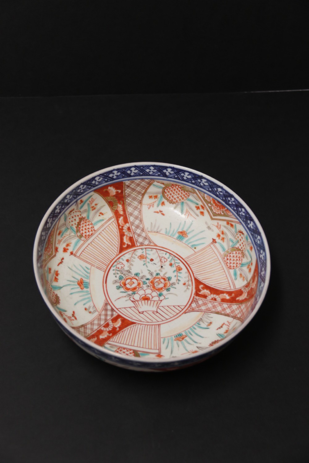 A round hand-painted Japanese porcelain bowl featuring underglaze blue geometric rim borders, iron-red floral panel motifs, and gilded details on a white background. View IX