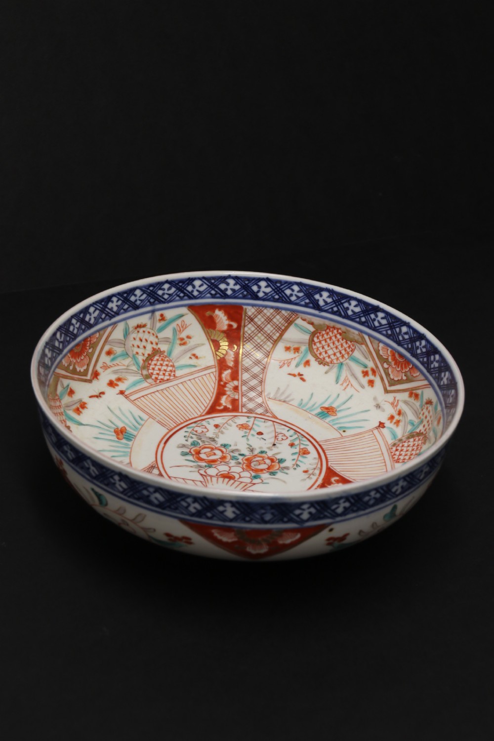 A round hand-painted Japanese porcelain bowl featuring underglaze blue geometric rim borders, iron-red floral panel motifs, and gilded details on a white background. View X