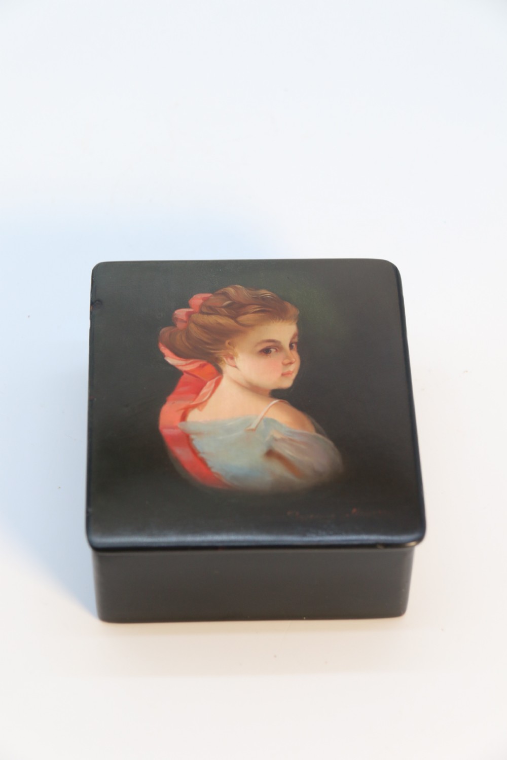 A square black lacquered box featuring a hand-painted portrait of a young woman with brown hair and a red ribbon. View III