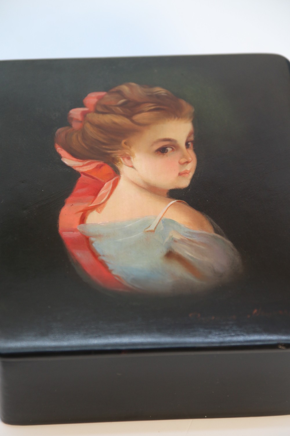 A square black lacquered box featuring a hand-painted portrait of a young woman with brown hair and a red ribbon. View IV