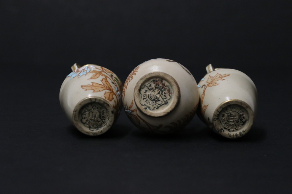 A set of three antique miniature cream ceramic vases featuring hand-painted floral motifs in blue, green, and brown with gilded highlights. View X