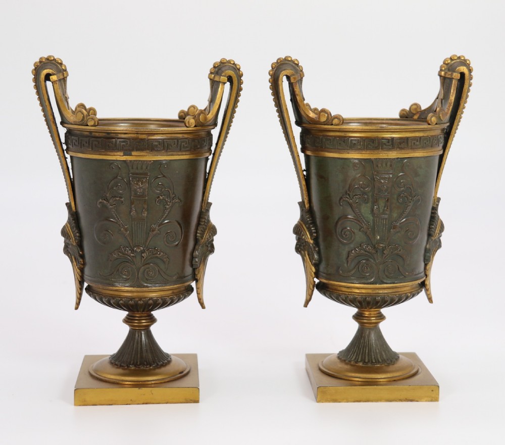 A pair of French Empire patinated and gilt bronze urns with Greek key friezes, scroll handles, and square bases.