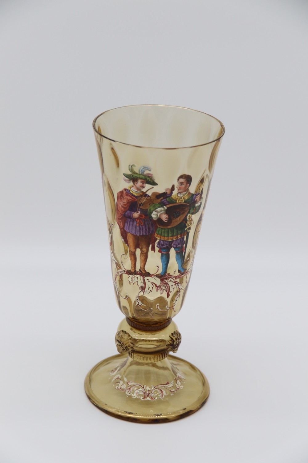 A pair of tall amber glass goblets featuring hand-painted enamelled figures of Renaissance musicians with scalloped rims and decorative knopped stems. View V