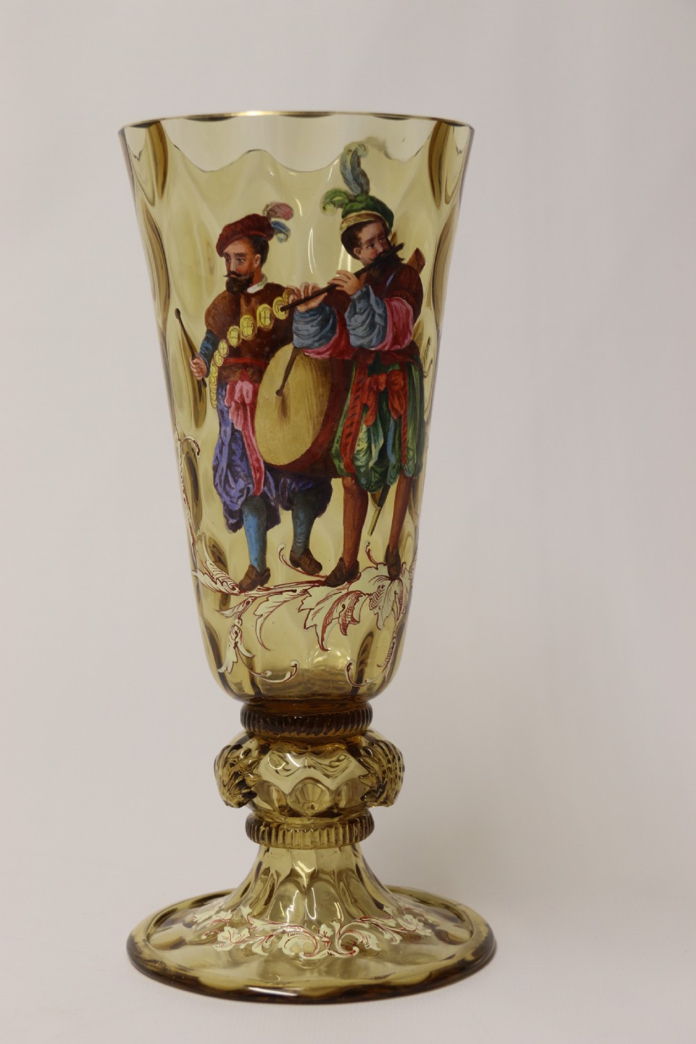 A pair of tall amber glass goblets featuring hand-painted enamelled figures of Renaissance musicians with scalloped rims and decorative knopped stems. View VI