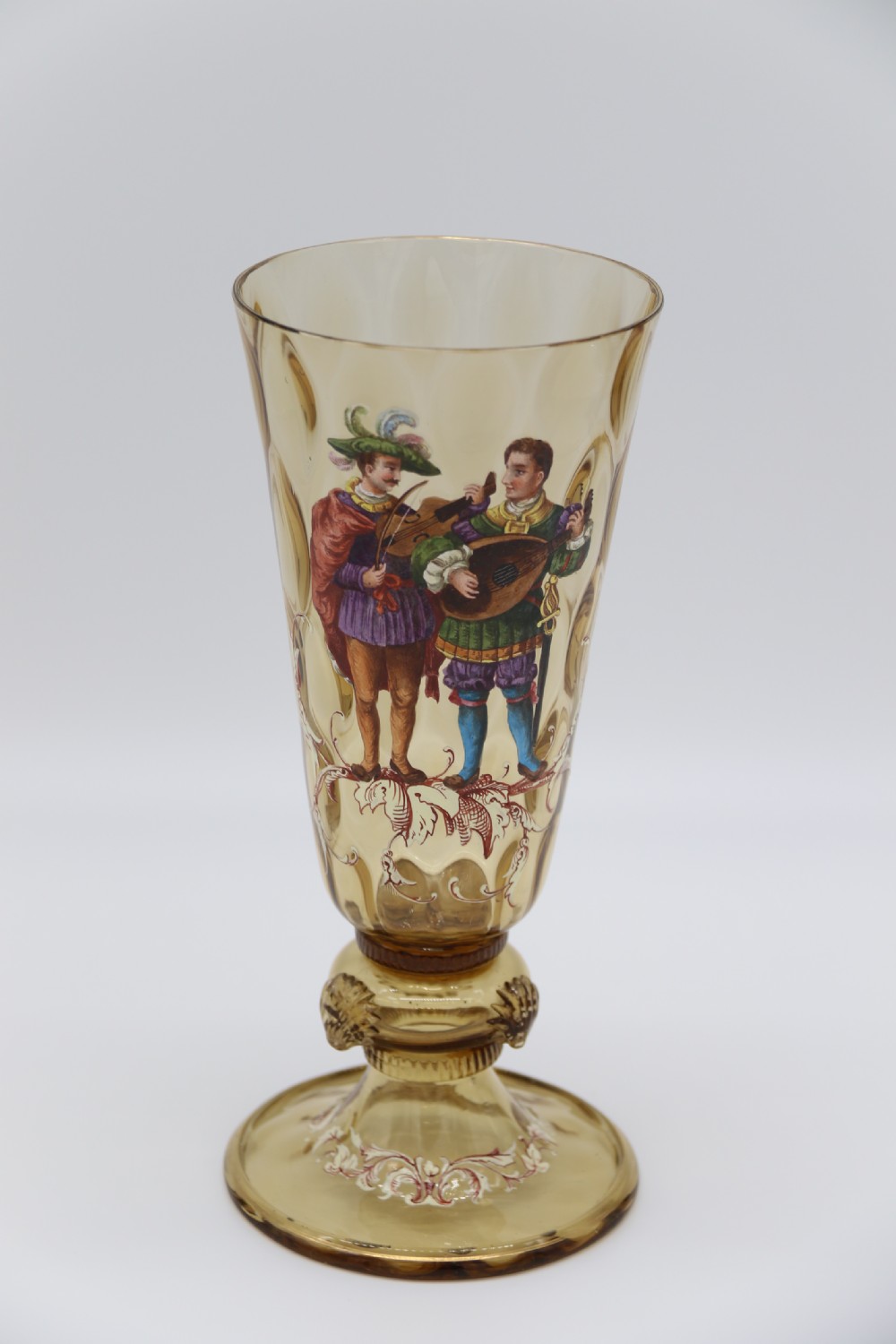 A pair of tall amber glass goblets featuring hand-painted enamelled figures of Renaissance musicians with scalloped rims and decorative knopped stems. View VII