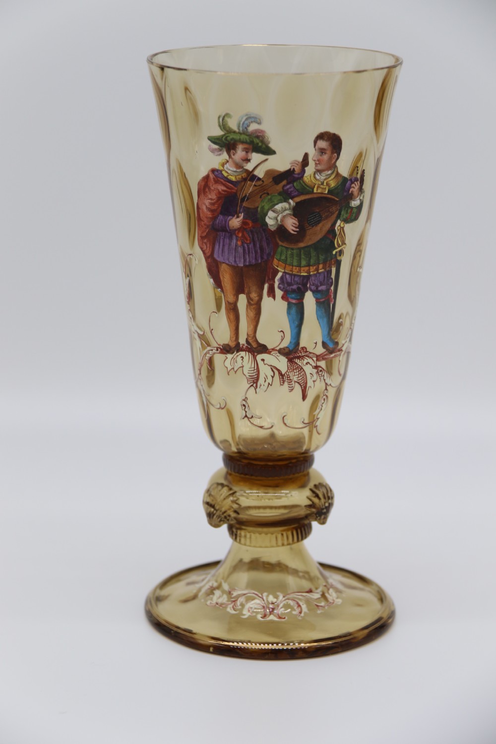 A pair of tall amber glass goblets featuring hand-painted enamelled figures of Renaissance musicians with scalloped rims and decorative knopped stems. View VIII