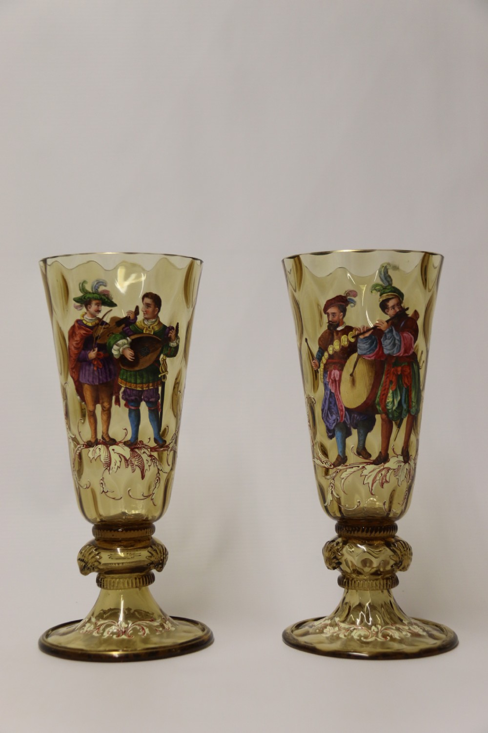 A pair of tall amber glass goblets featuring hand-painted enamelled figures of Renaissance musicians with scalloped rims and decorative knopped stems. View IX