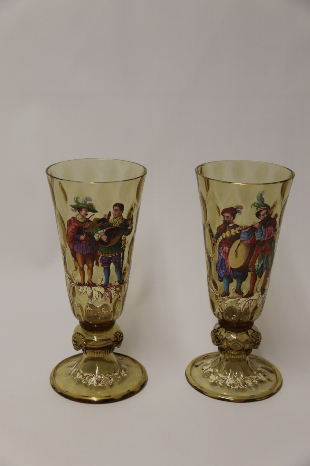 A pair of tall amber glass goblets featuring hand-painted enamelled figures of Renaissance musicians with scalloped rims and decorative knopped stems. View X