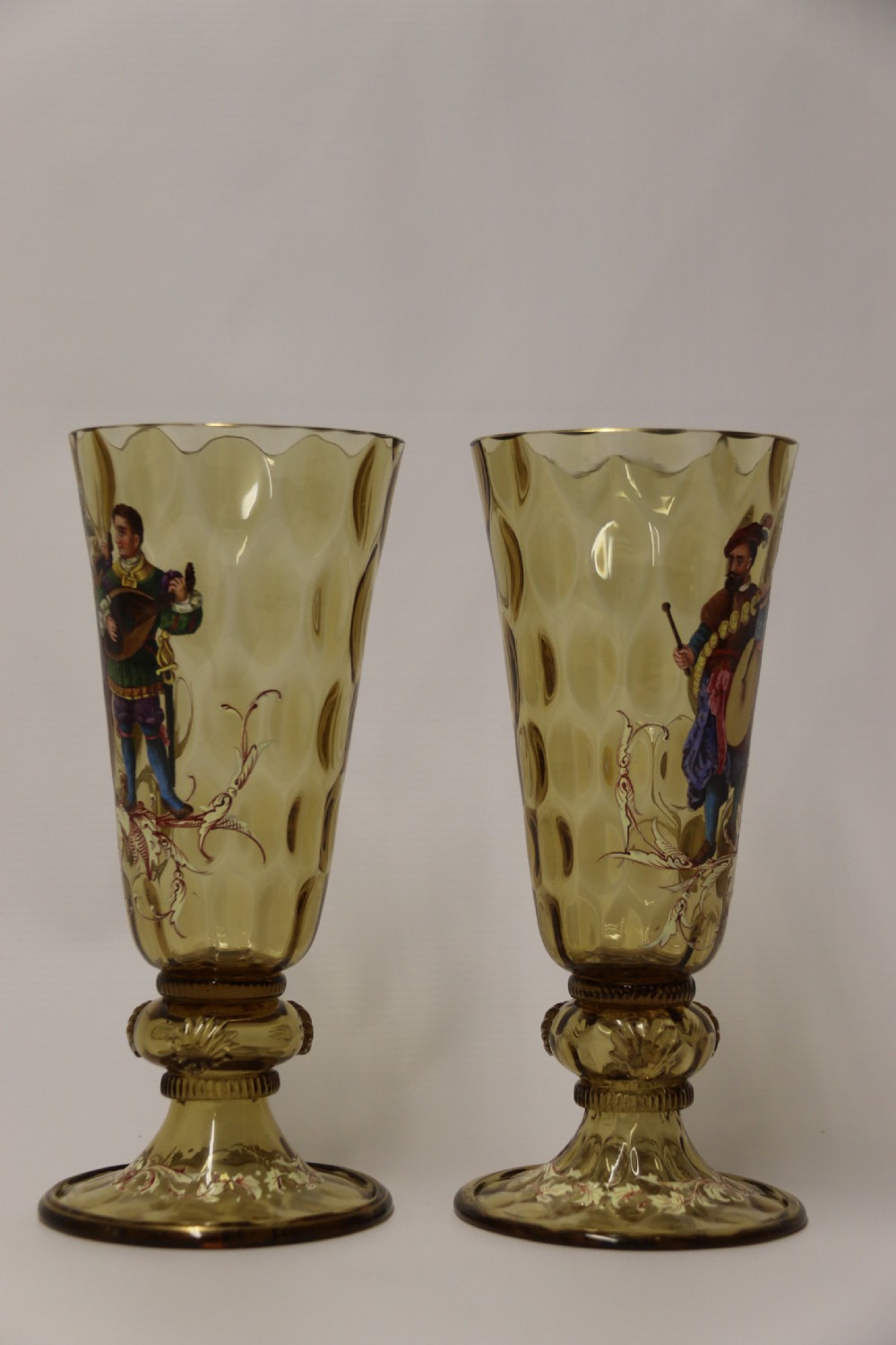 A pair of tall amber glass goblets featuring hand-painted enamelled figures of Renaissance musicians with scalloped rims and decorative knopped stems. View XI