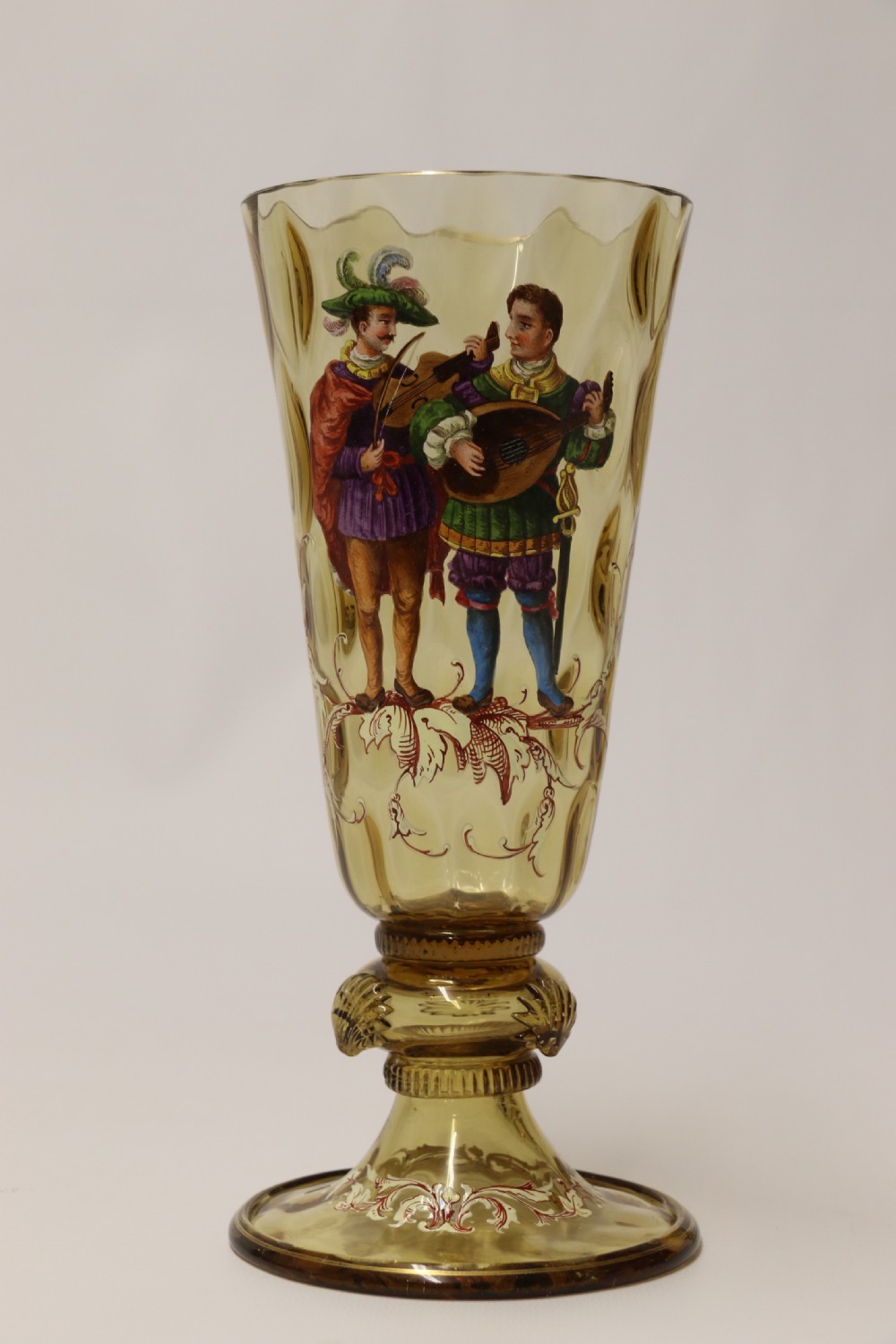 A pair of tall amber glass goblets featuring hand-painted enamelled figures of Renaissance musicians with scalloped rims and decorative knopped stems. View XVII