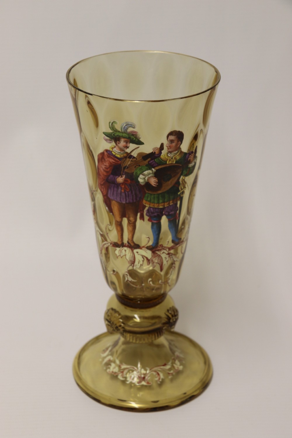 A pair of tall amber glass goblets featuring hand-painted enamelled figures of Renaissance musicians with scalloped rims and decorative knopped stems. View XXV