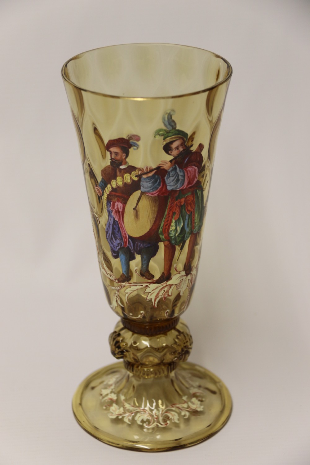 A pair of tall amber glass goblets featuring hand-painted enamelled figures of Renaissance musicians with scalloped rims and decorative knopped stems. View XXVI