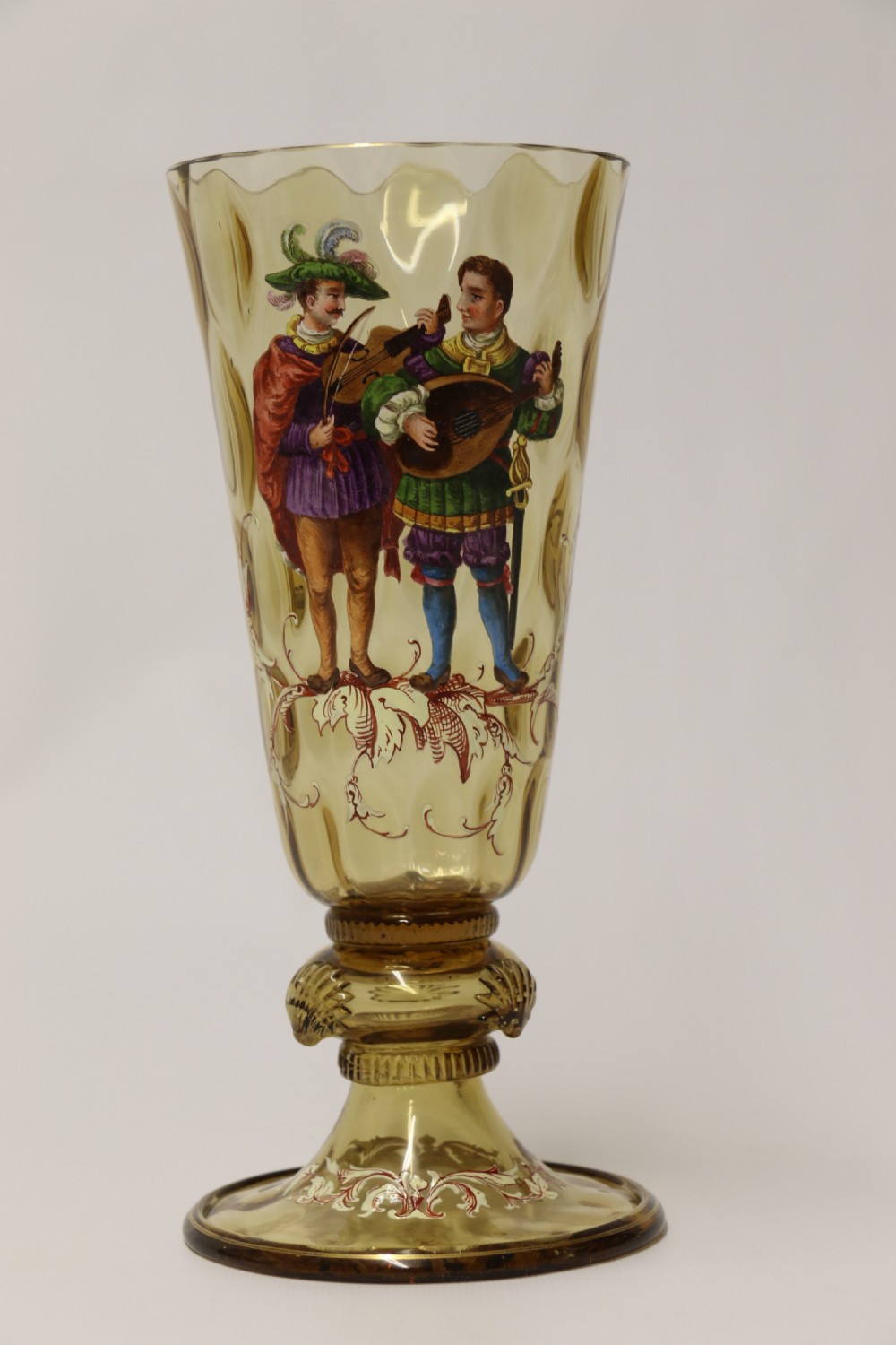 A pair of tall amber glass goblets featuring hand-painted enamelled figures of Renaissance musicians with scalloped rims and decorative knopped stems. View XXXI