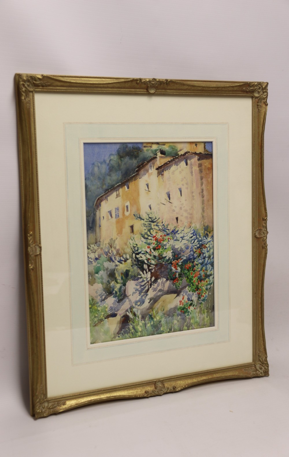 A framed 1920s watercolour painting depicting a sunlit Mediterranean building behind foliage, set within an ornate gilded frame.