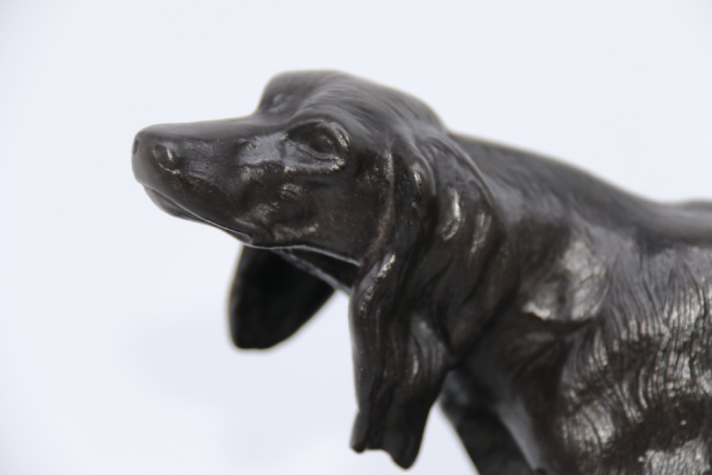 A black cast iron sculpture depicting a hunting dog walking across a naturalistic earth base. View III