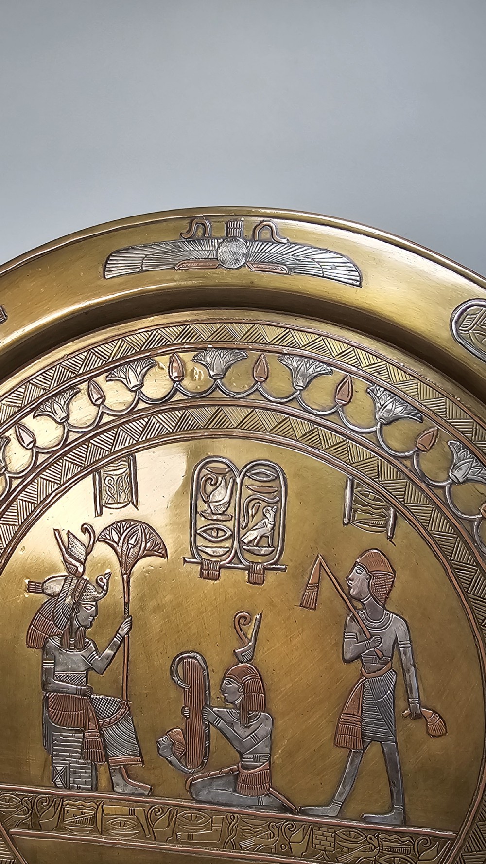A circular brass charger featuring inlaid silver and copper depiction of Ancient Egyptian figures, hieroglyphs, and winged sun disc motifs. View V