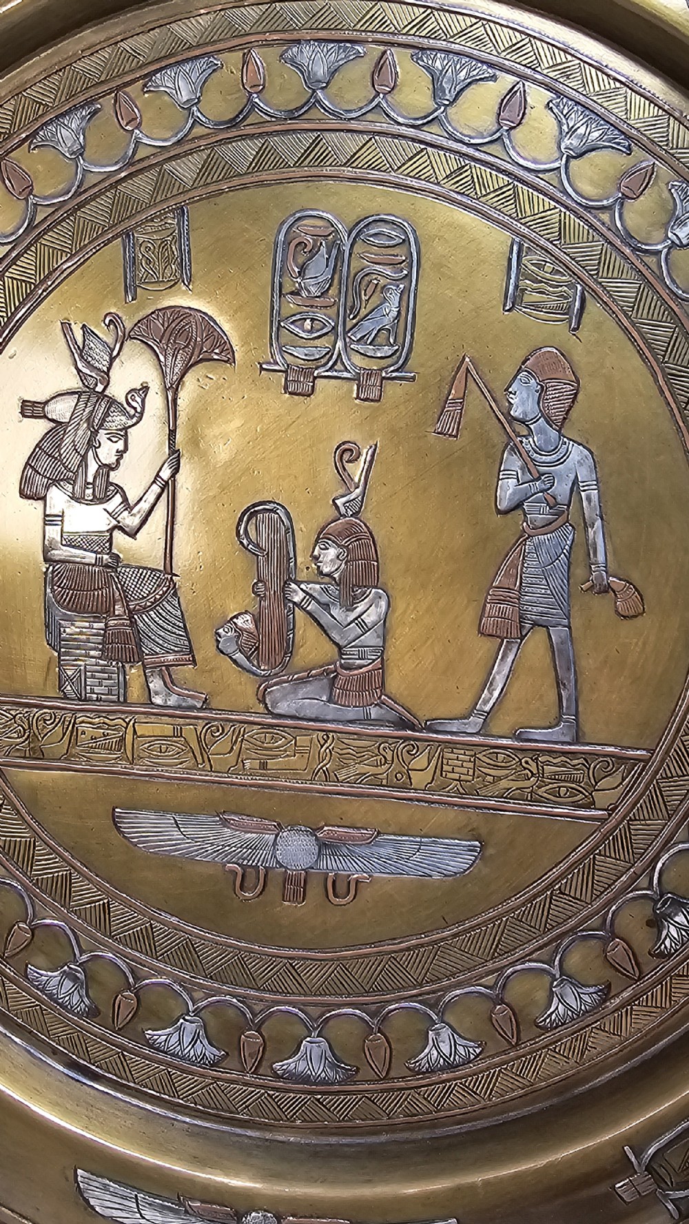 A circular brass charger featuring inlaid silver and copper depiction of Ancient Egyptian figures, hieroglyphs, and winged sun disc motifs. View VII