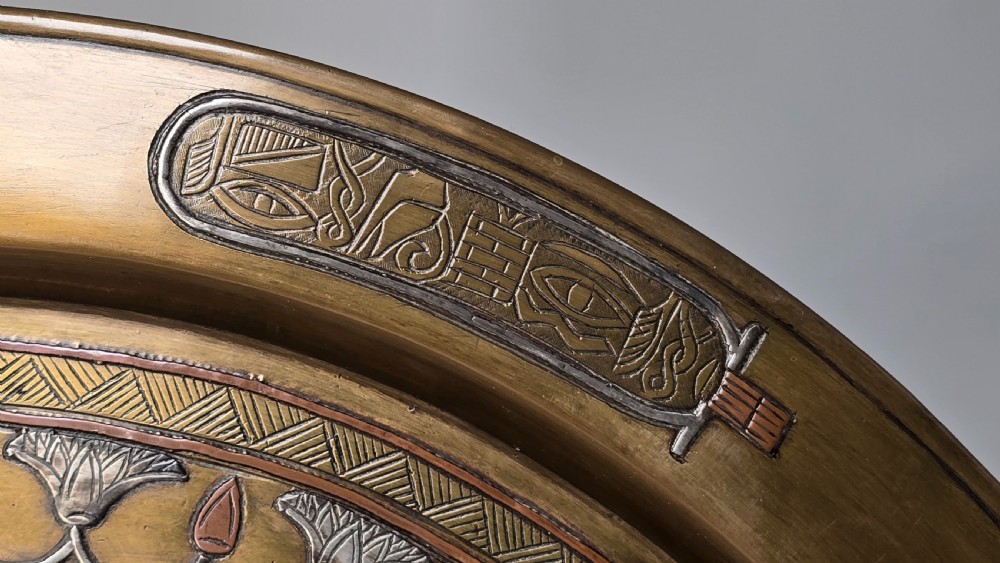 A circular brass charger featuring inlaid silver and copper depiction of Ancient Egyptian figures, hieroglyphs, and winged sun disc motifs. View XV