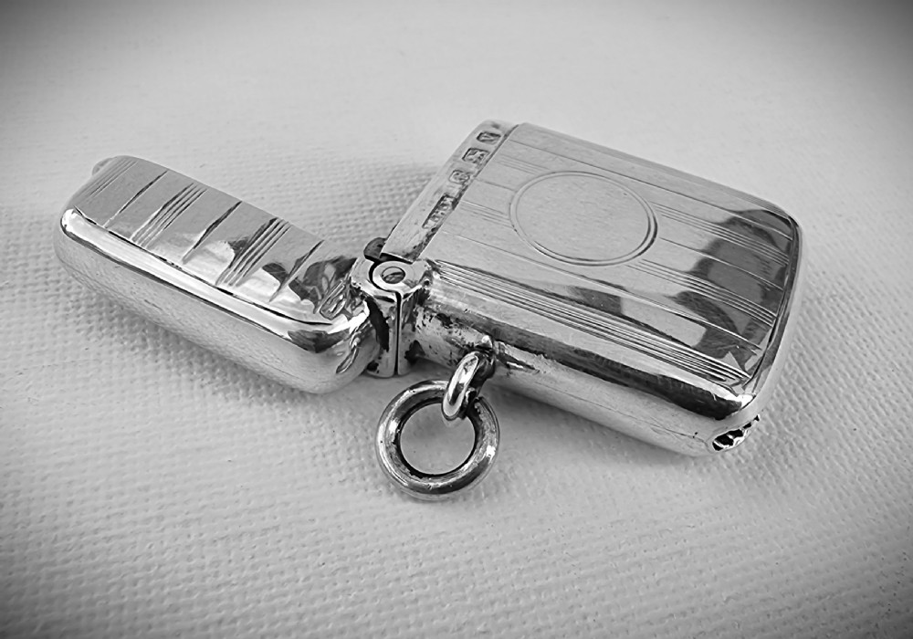 An open antique sterling silver vesta case featuring linear engine-turned engraving, a central circular cartouche, British hallmarks, and a suspension ring.