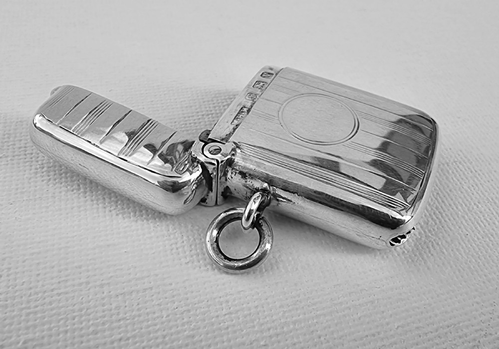 An open antique sterling silver vesta case featuring linear engine-turned engraving, a central circular cartouche, British hallmarks, and a suspension ring. View IV