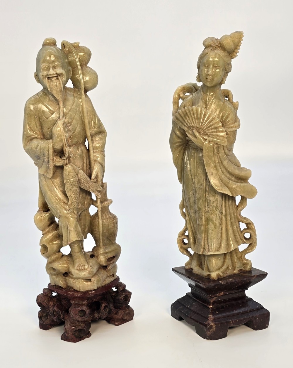A pair of carved pale green stone Chinese figures representing a fisherman with a fish and a maiden with a fan, displayed on dark wood bases.