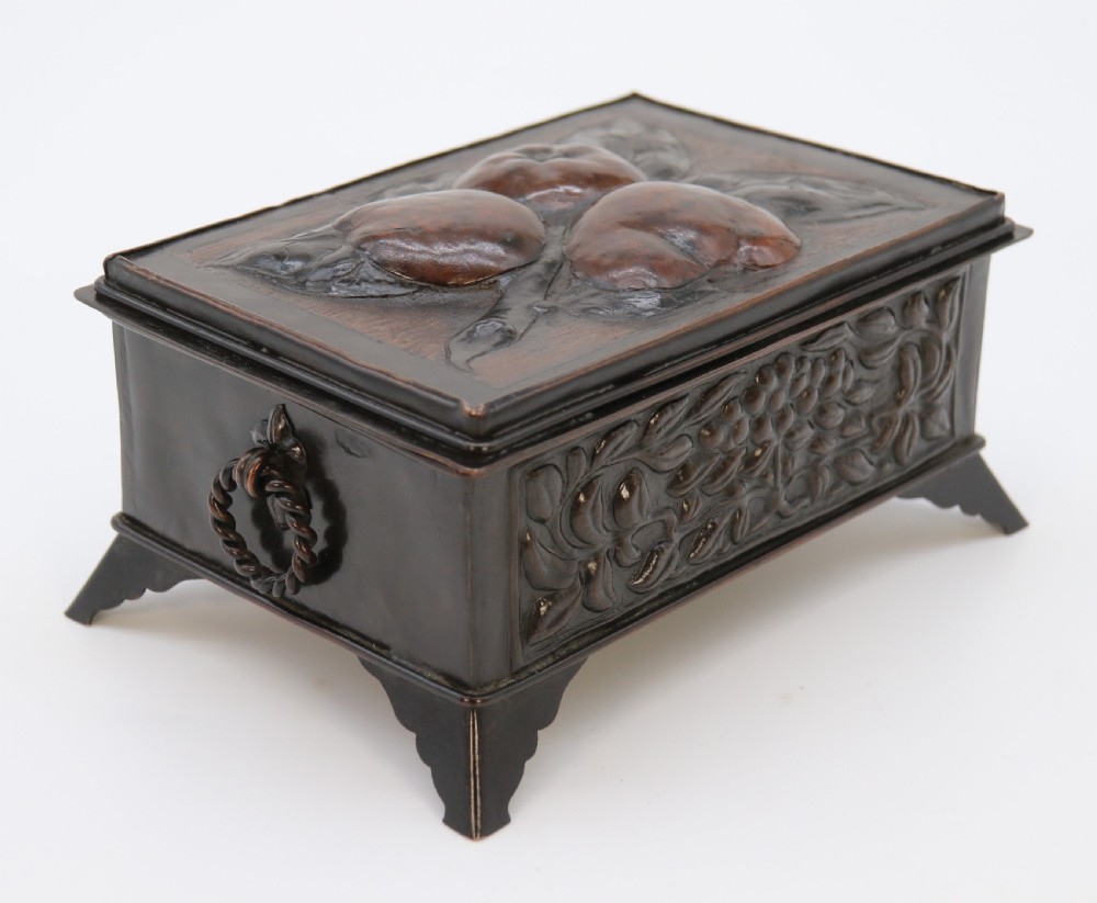 A rectangular dark patinated copper box featuring repousse fruit motifs on the lid, stylized foliage on the front face, bracket feet, and side drop ring handles. View III