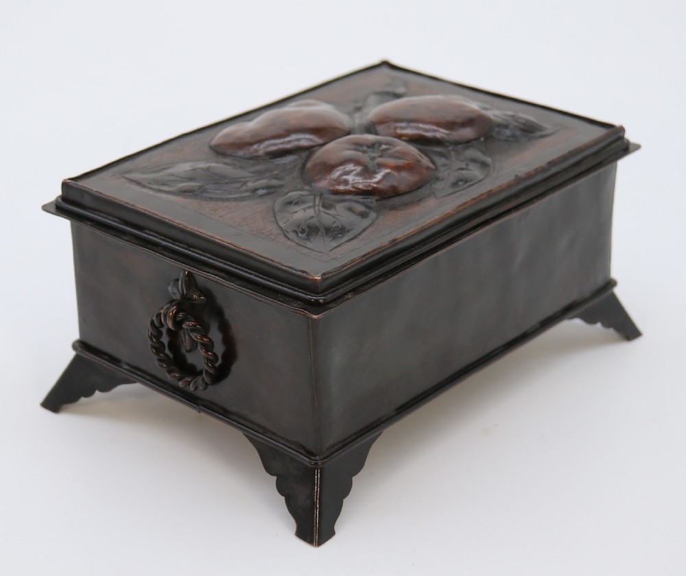 A rectangular dark patinated copper box featuring repousse fruit motifs on the lid, stylized foliage on the front face, bracket feet, and side drop ring handles. View V