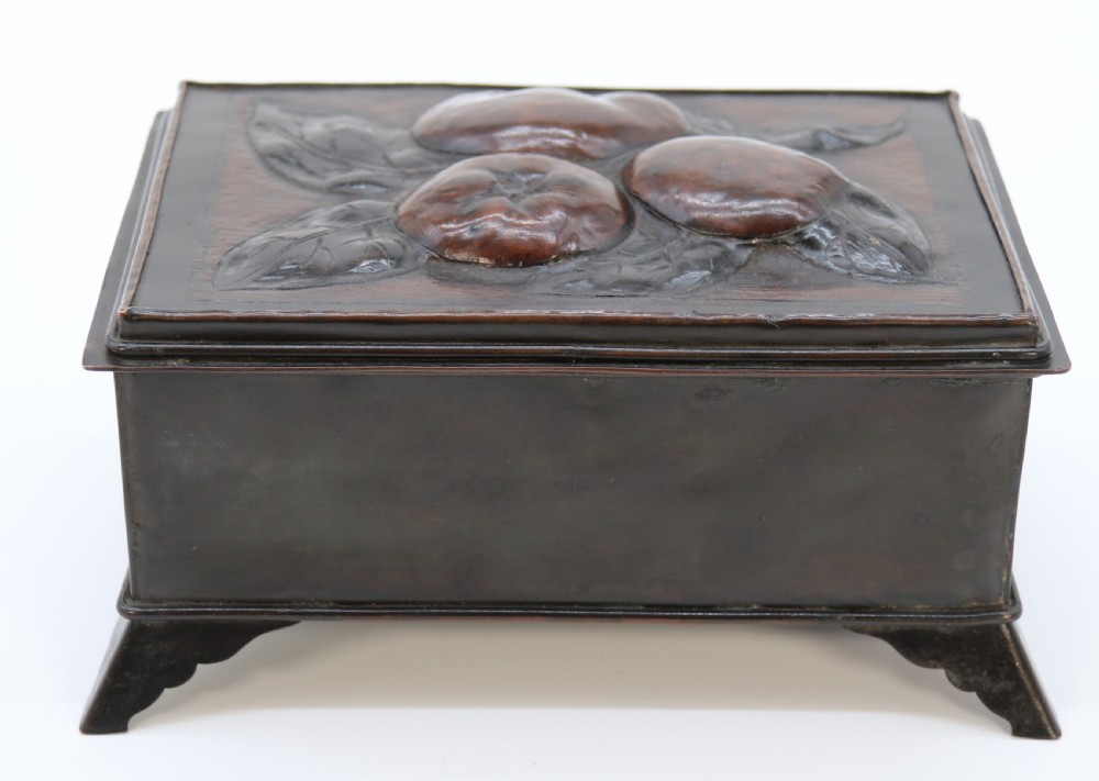 A rectangular dark patinated copper box featuring repousse fruit motifs on the lid, stylized foliage on the front face, bracket feet, and side drop ring handles. View VI