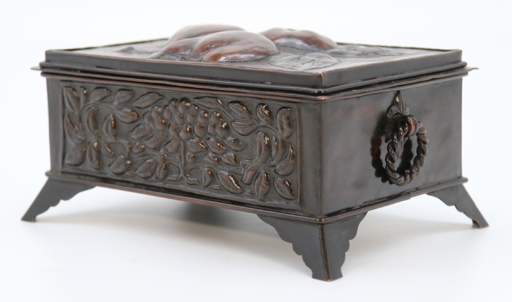 A rectangular dark patinated copper box featuring repousse fruit motifs on the lid, stylized foliage on the front face, bracket feet, and side drop ring handles. View VIII
