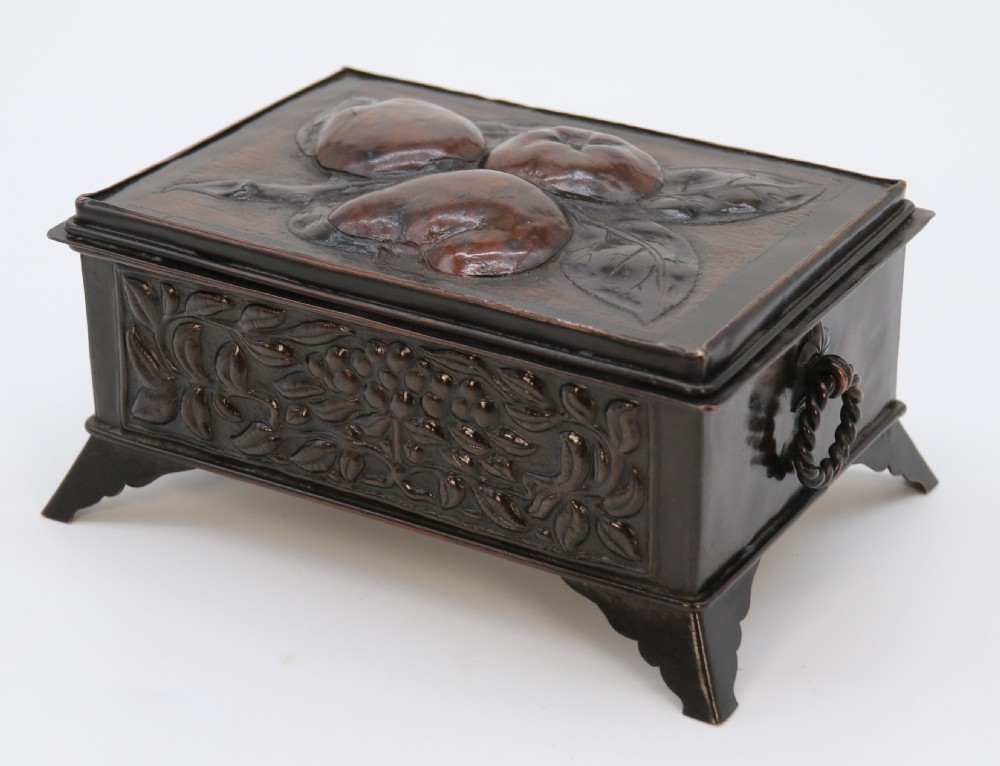 A rectangular dark patinated copper box featuring repousse fruit motifs on the lid, stylized foliage on the front face, bracket feet, and side drop ring handles. View X