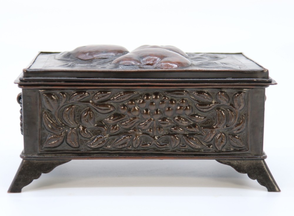A rectangular dark patinated copper box featuring repousse fruit motifs on the lid, stylized foliage on the front face, bracket feet, and side drop ring handles. View XI