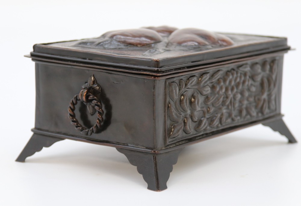 A rectangular dark patinated copper box featuring repousse fruit motifs on the lid, stylized foliage on the front face, bracket feet, and side drop ring handles. View XIII