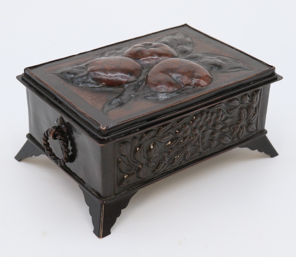 A rectangular dark patinated copper box featuring repousse fruit motifs on the lid, stylized foliage on the front face, bracket feet, and side drop ring handles. View XIX