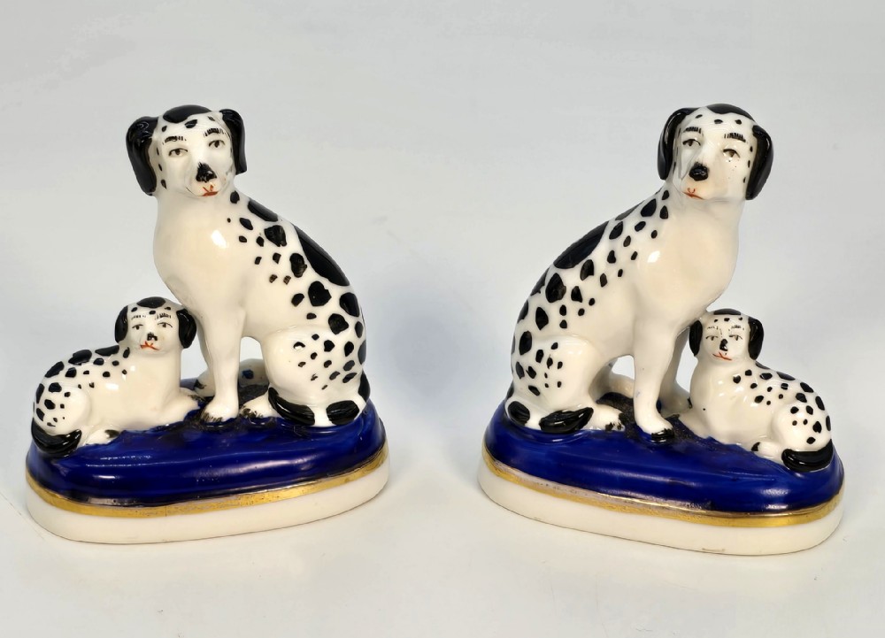 A pair of antique porcelain figurines depicting seated black-and-white spotted Dalmatian dogs with puppies on cobalt blue bases with gilt trim.