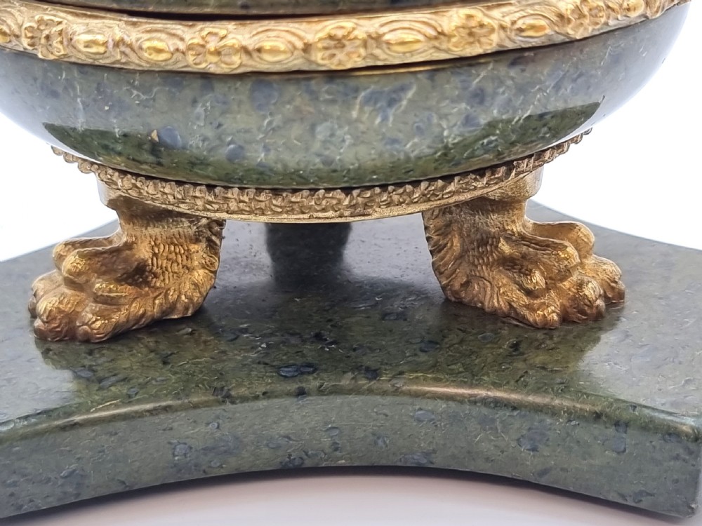 A French Empire style green marble inkstand featuring a central urn-shaped vessel with gilt bronze floral borders and a decorative finial lid, supported by three lion paw feet on a curved marble base. View VI