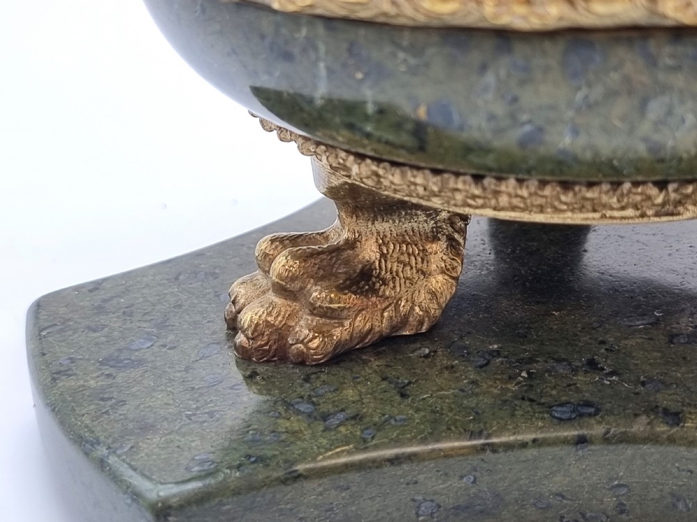 A French Empire style green marble inkstand featuring a central urn-shaped vessel with gilt bronze floral borders and a decorative finial lid, supported by three lion paw feet on a curved marble base. View XVI